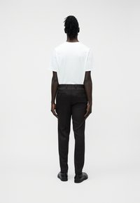 Άτομο με σκούρα πλεγμένα μαλλιά στέκεται γυρισμένο προς τα πίσω, φοράει λευκό κοντομάνικο πουκάμισο, μαύρο παντελόνι και μαύρα παπούτσια μπροστά σε λευκό φόντο.