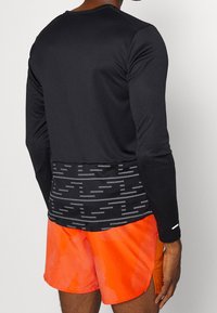 Rückansicht einer Person, die ein schwarzes Langarmshirt mit grauem horizontalem Streifenmuster und leuchtend orangefarbenen Sportshorts trägt.