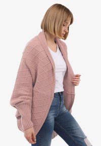 Frau mit kurzen blonden Haaren, die eine strukturierte rosa Strickjacke über einem weißen T-Shirt und blauen Jeans trägt. Sie schaut nach unten und hält die Strickjacke geöffnet.