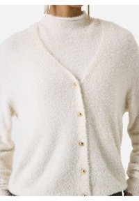 Vrouw in een crèmekleurige fuzzy cardigan die asymmetrisch is dichtgeknoopt over een bijpassende fuzzy coltrui, met gouden hangende oorbellen zichtbaar.
