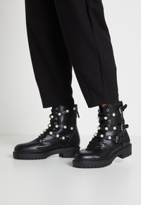 Stivali da combattimento in pelle nera con cinturini e fibbie decorati con perle, indossati con pantaloni neri a sigaretta su un pavimento bianco.