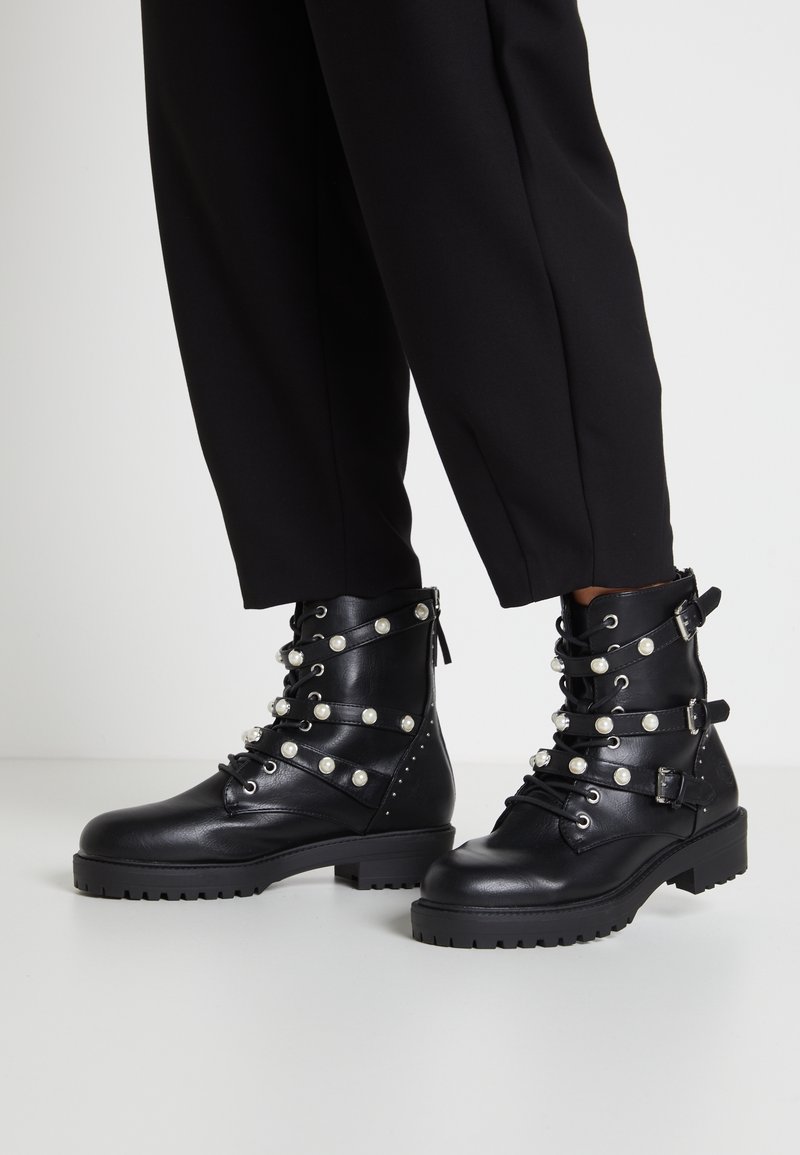 Stivali da combattimento in pelle nera con cinturini e fibbie decorati con perle, indossati con pantaloni neri a sigaretta su un pavimento bianco.