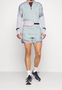 Camiseta atlética de color azul claro con acentos morados, combinada con shorts a juego que presentan un patrón floral. También incluye una riñonera gris y zapatos negros.