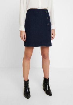 Personne portant une jupe bleu marine avec des boutons latéraux, un chemisier blanc à manches longues et des bottines noires, se tenant devant un fond uni.