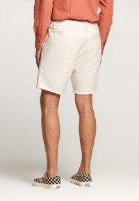 Bermudas de algodón beige con un ajuste clásico y bolsillos laterales, combinadas con zapatos de lona negros y blancos a cuadros. La tela parece suave.