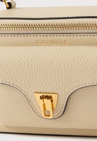 Beżowa skórzana torebka o fakturowanej powierzchni, złotych elementach i charakterystycznym trójkątnym zapięciu. Wyposażona w zamek błyskawiczny i zdobienie z logo.