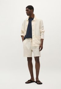 Linnedragt i creme med knaplukning, parret med en navy t-shirt og creme shorts. Sorte flip-flops fuldender looket.