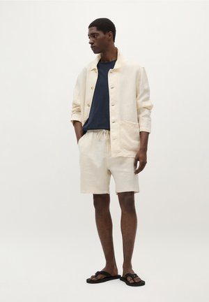 Chaqueta de lino en crema con botones al frente, combinada con una camiseta navy y pantalones cortos en crema. Chanclas negras completan el look.