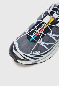 Pelēkie sporta apavi ar tīkla augšdaļu, gumijas zoli un baltām detaļām. Iespējas ietver elastīgās šņores, ar zīmolu apzīmētu valni un pastiprinātu pirkstu daļu.