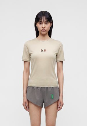 Une femme porte un t-shirt beige avec logo et un short de course gris sur un fond clair uni.