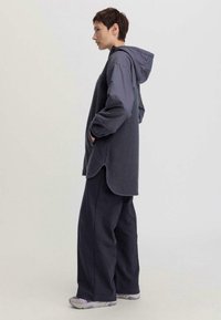 Giacca a felpa grigia con cappuccio, maniche lunghe e tasche laterali, abbinata a pantaloni larghi grigio scuro e sneakers grigio chiaro.