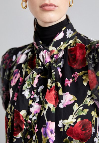 Floral blouse in black fabric with red, pink, and green patterns. Features a bow tie at the collar and button details along the front.