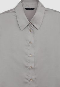 Camicia button-up grigia chiara con colletto a punta, cinque bottoni beige e un'etichetta "CALLIOPE" all'interno del colletto.