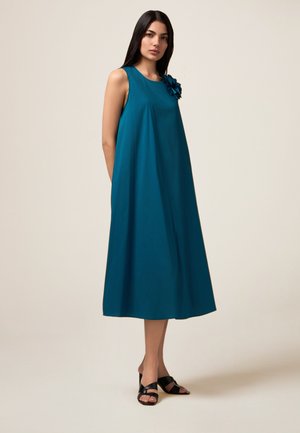 Türkisfarbenes, ärmelloses Kleid mit lockerer Passform, das einen floralem Akzent an der linken Schulter hat. Glattes Material und knielange Länge. Schwarze, hochhackige Sandalen.