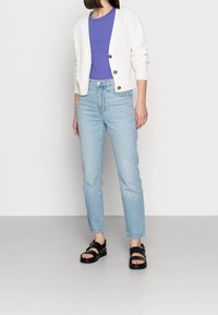 Valkoinen ribattu neuletakki, violetti T-paita, vaaleansiniset farkut ja mustat sandaalit. Neuletakissa on napit; farkuissa on suora leikkaus.