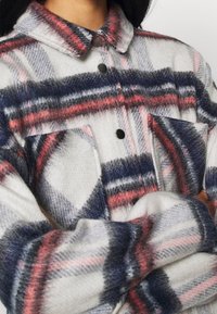 Een flanellen shirt in grijs, wit en marineblauw met rode accenten. Voorzien van een kraag, zwarte knopen en voorzakken. Zachte textuur.