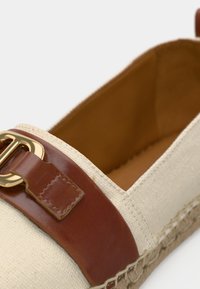 Μπεζ πάνινο παπούτσι τύπου slip-on με καφέ δερμάτινο λουράκι και λεπτομέρεια με χρυσή αγκράφα. Φυσική σόλα από ίνες με υφασμένη υφή.