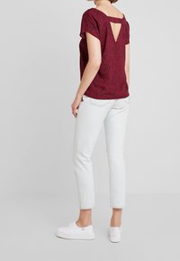 Burgunderfarvet kortærmet top med et plettet mønster, scoop ryg med en nøglehulskæring, parret med lyseblå jeans og hvide sneakers.