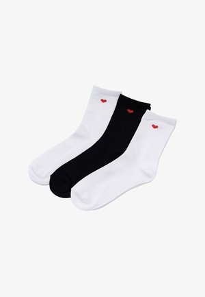 Trzy skarpetki do kostki, dwie białe i jedna czarna, każda z małym czerwonym sercem blisko górnej krawędzi, ułożone obok siebie.