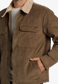 Chaqueta de pana marrón con un suave collar color beige, dos bolsillos en el pecho y bolsillos laterales. La tela tiene una textura de líneas verticales y un cierre de cremallera.