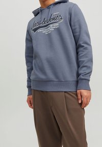 Szary bawełniany hoodie z sznurkiem, z białym logo na przodzie oraz kieszenią kangurką, zestawiony z luźnymi brązowymi spodniami.