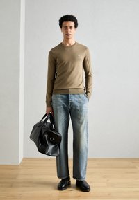 Bruine gebreide trui met ronde hals, blauwe denim jeans en zwarte leren sporttas. Model staat op een houten vloer tegen een lichte achtergrond.