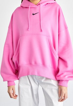 Persona que lleva una sudadera Nike de gran tamaño de color rosa vivo con bolsillos delanteros y pantalones blancos sobre un fondo sencillo.