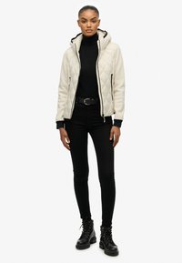 Biała pikowany kurtka z kapturem z czarnymi akcentami, z zamykanymi kieszeniami, noszona na czarnym golfie i czarnych wąskich dżinsach, w połączeniu z czarnymi botkami.