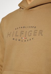Brown hooded sweatshirt with a front logo that reads "ESTABLISHED HILFIGER MCMLXXXV." Features a soft texture and kangaroo pocket.