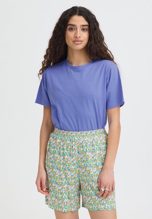 Młoda kobieta z długimi kręconymi włosami, ubrana w gładki fioletowy t-shirt i szorty w kwiatowy wzór na jasnym tle.