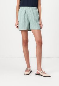 Shorts verts clairs avec un motif zigzag texturé, dotés d'une taille élastique. Associés à des chaussures à talon ouvert beiges.