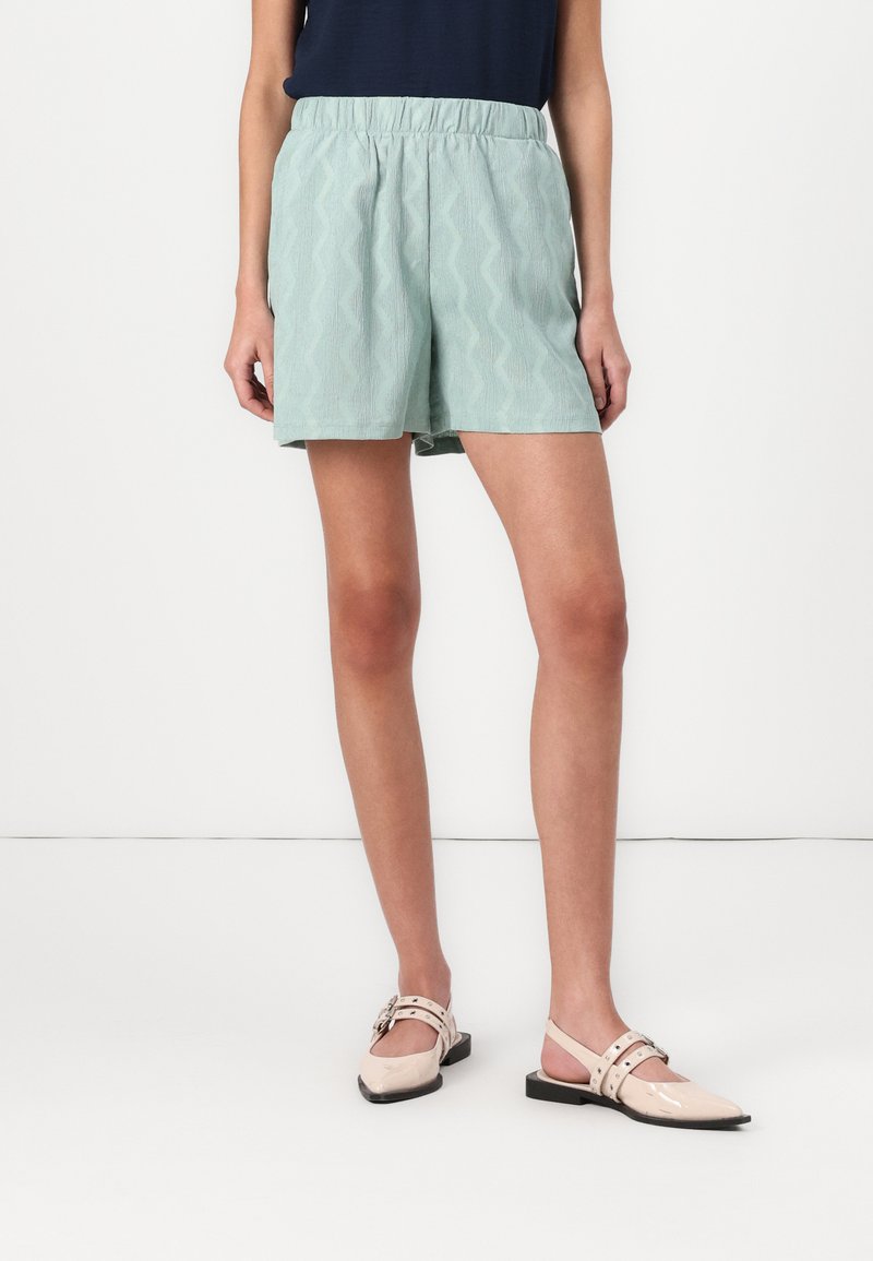 Shorts verts clairs avec un motif zigzag texturé, dotés d'une taille élastique. Associés à des chaussures à talon ouvert beiges.