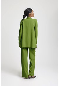 Kvinna med flätat hår iklädd en limegrön blazer och matchande vida byxor, stående på sidan mot en vit bakgrund.