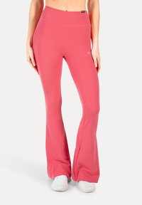 Pantaloni da allenamento a campana di un rosa chiaro, realizzati in tessuto elasticizzato, con una vita alta e una texture liscia e lucida. Abbinati a scarpe da ginnastica bianche.