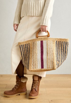 Pleciona torba ze słomy z drewnianą rączką, ozdobiona poziomymi paskami w kolorach beżowym, białym, niebieskim i bordo. Modelka ma na sobie brązowe buty ocieplane futerkiem.