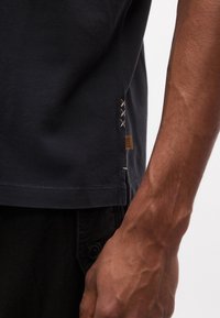 Κοντινή λήψη του χεριού και του κορμού ενός ατόμου που φοράει σκούρο κοντομάνικο πουκάμισο με μπεζ σταυρωτές ραφές και μια μικρή καφέ ετικέτα κοντά στο τελείωμα.