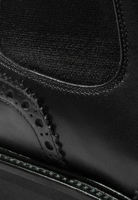Botín de cuero negro con superficie lisa, que cuenta con tejido texturizado, detalles decorativos de brogue y una suela negra resistente.