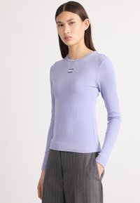 Langarm-Rippoberteil in hellem Lavendel, mit Rundhalsausschnitt und kleinem dekorativem Logo in der Mitte. Kombiniert mit nahtlosen Streifenhosen.