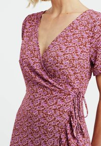 Vestido de estampado floral morado con fondo marrón, escote en V, mangas cortas y detalle de lazo en el costado. Tejido suave con un acabado ligeramente texturizado.