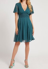 Nainen, joka käyttää turkoosia polviin ulottuvaa pilkullista mekkoa, jossa on lyhyet liehuvat hihat, ja avonaiset turkoosit korkokengät, seisoo yksinkertaista taustaa vasten.