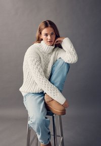 Jeune femme portant un pull à col roulé en maille torsadée blanche, un jean bleu clair et des bottes beiges doublées de fourrure, assise sur un tabouret devant un fond gris.