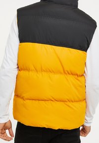 Veste sans manches matelassée avec un panneau supérieur noir et un panneau inférieur jaune vif, dotée d'une texture lisse et de détails de couture visibles.