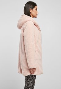 Cappotto con cappuccio fucsia, morbido e con una vestibilità ampia, superficie testurizzata e tasche laterali. Indossato sopra pantaloni neri a fantasia.