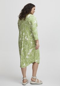 Vestido verde con un estampado, que presenta un corte holgado, mangas de tres cuartos y una cintura fruncida. Combinado con sandalias blancas de plataforma.