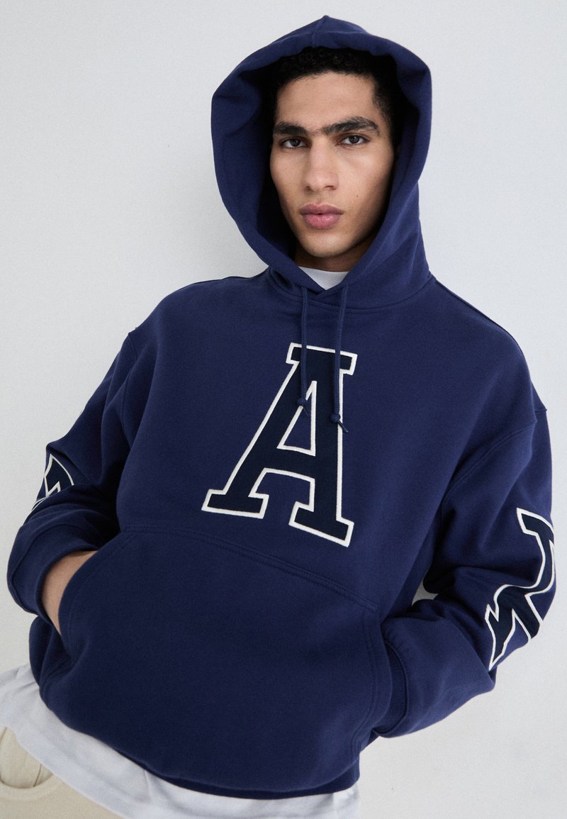 Navyblaue Kapuzenjacke aus weichem Material mit einem großen weißen "A"-Logo auf der Vorderseite und dekorativen Mustern an den Ärmeln. Verstellbare Kapuze.