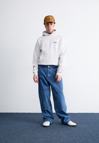 Hanorac gri cu buzunar frontal, blugi evazați albaștri cu cusături aplicate și adidași albi. Căciulă maro cu logo. Fundal uniform.