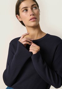 Marine crew-neck sweater med lange ærmer, ribbet tekstur og tommelhuller, båret med en afslappet pasform. Stoffet fremstår blødt og let.