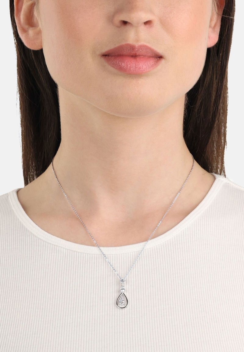 Kvinna som bär ett silverhalsband med en tårformad hänge med en central klar ädelsten, visad mot en vit rippad överdel.