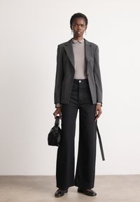 Blazer ajusté gris sur un haut gris clair, associé à un pantalon noir à jambe large avec une texture subtile et des embellissements. Sac à main noir.