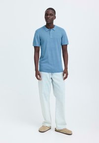 Polo à manches courtes bleu clair avec col, patte de boutonnage à deux boutons et ourlet côtelé. Associé à un jean en denim clair ample et des chaussons beige.